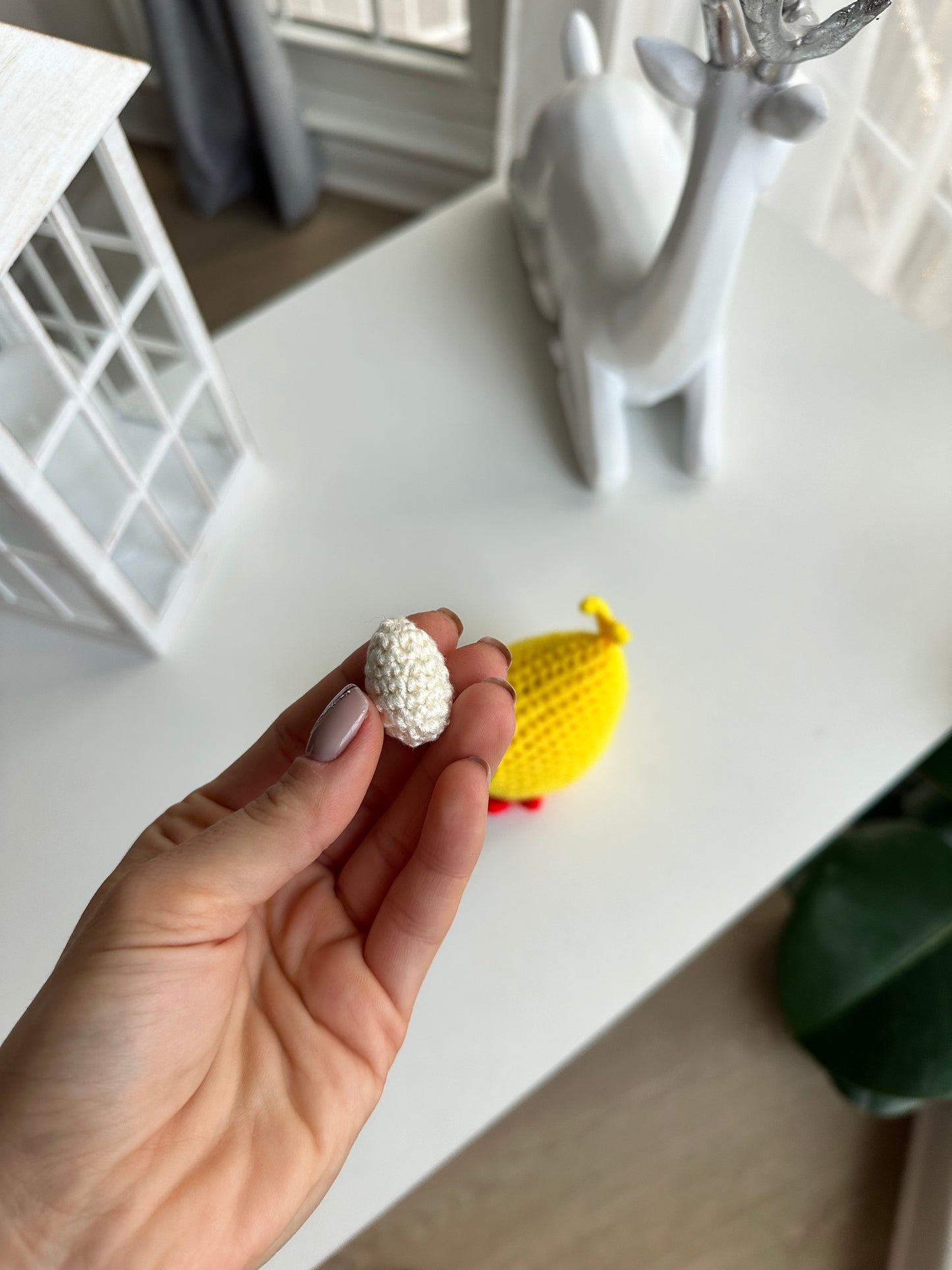 Hand holding a small white egg next to a yellow chicken keychain on a white surface with a blurred background