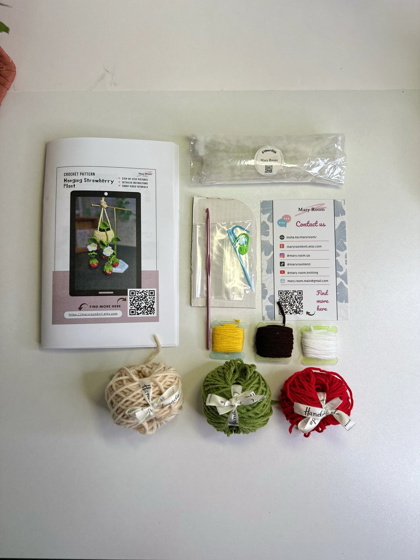 Flat lay showing red, green, cream yarn balls, cardboard, fiberfill, and crochet hook for strawberry kit