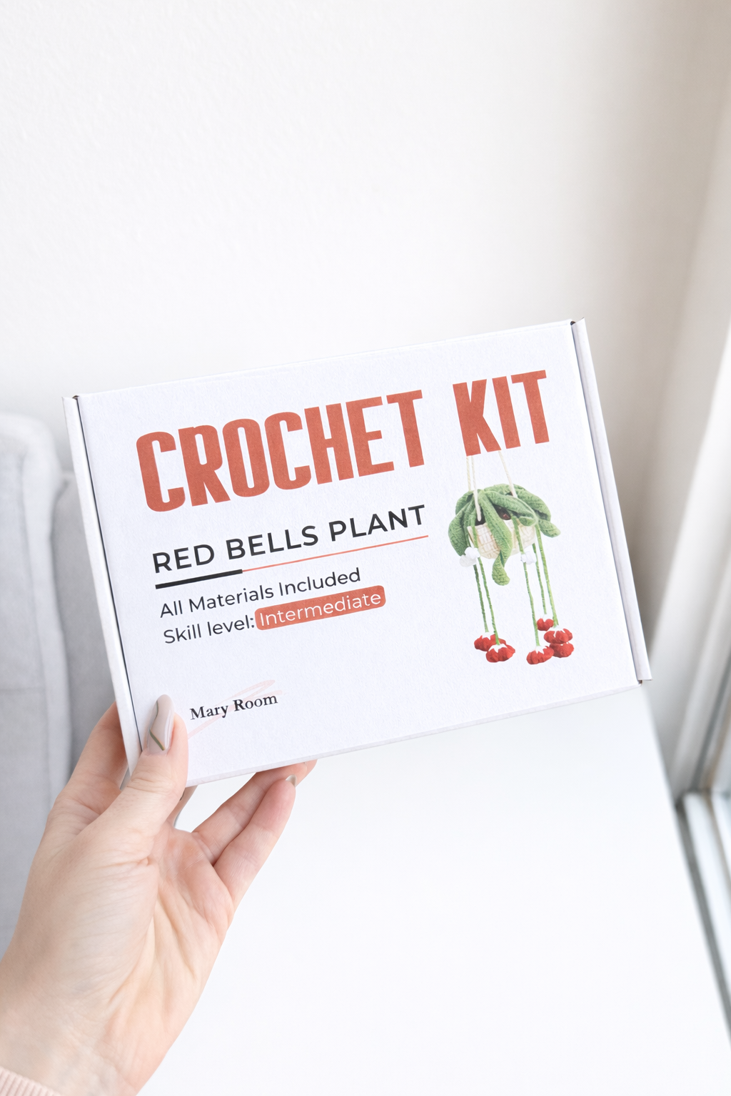 Holding the product box labeled DIY Crochet Kit Red Bells Plant, white minimal background.