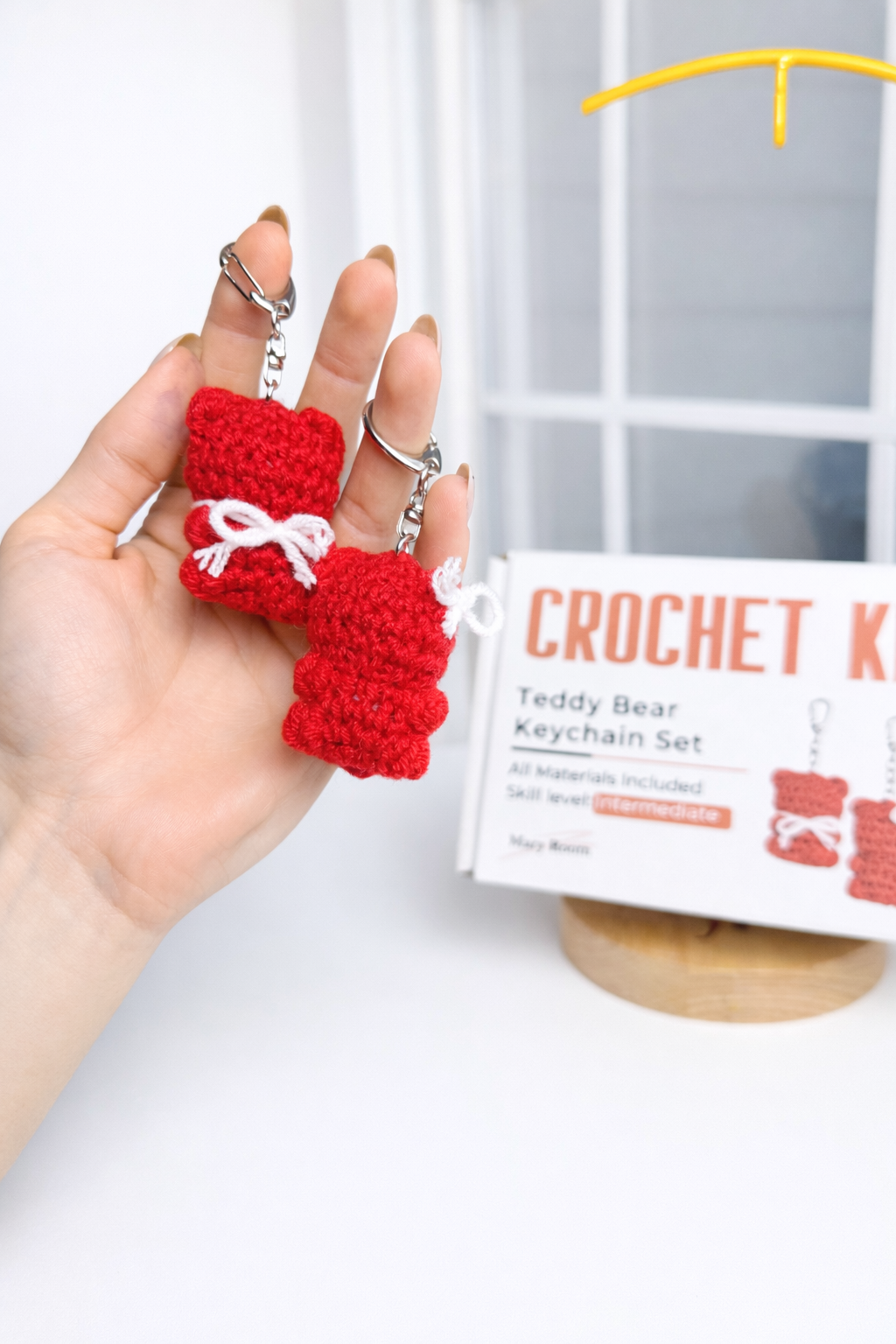 Holding one finished red teddy bear keychain with kit packaging in the background – DIY Crochet Kit for Valentine’s Day (Kissing Bears)