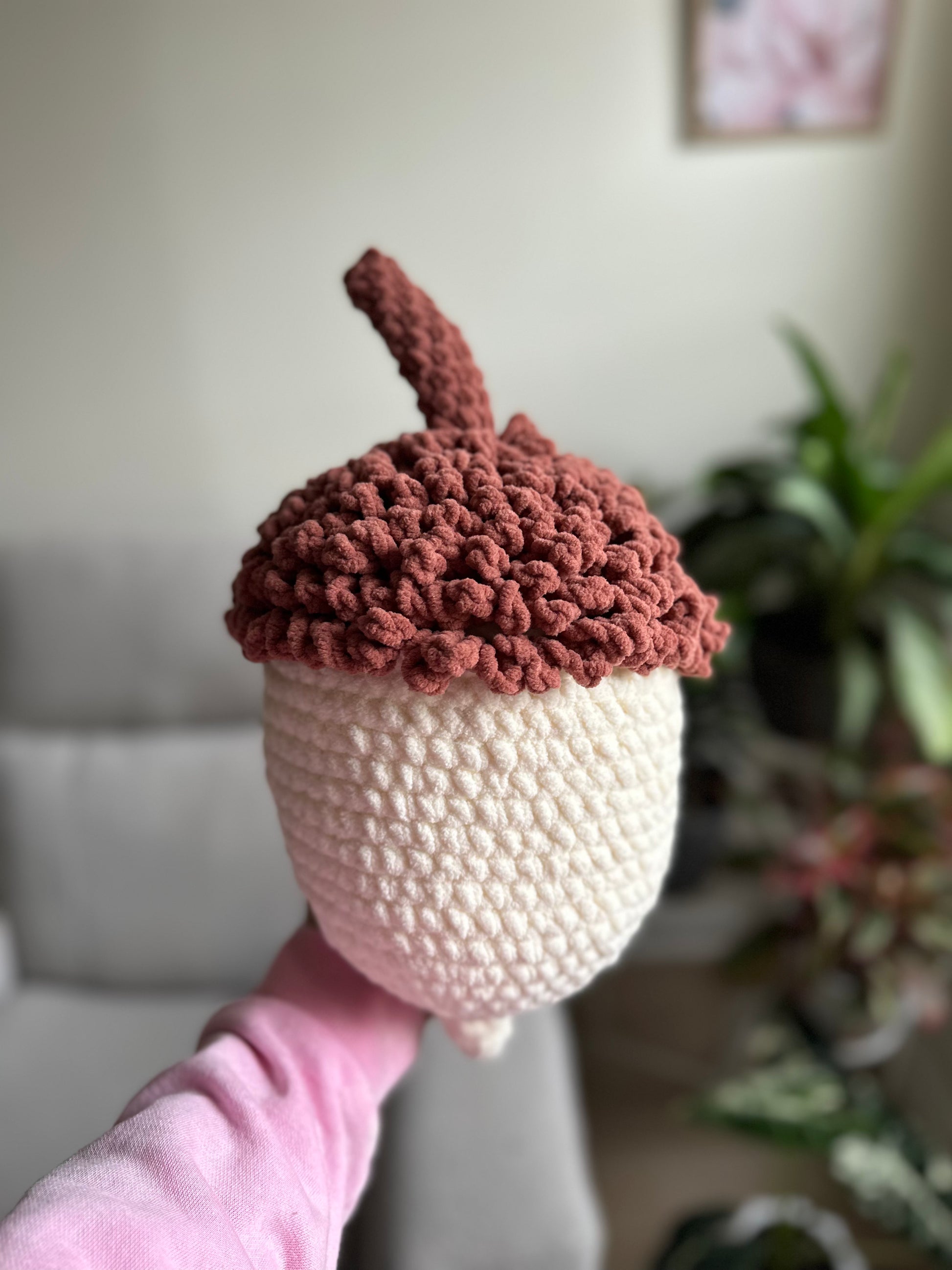A crocheted acorn-shaped plush toy or decorative item held up by a person's hand. The top half is a dusty rose-brown textured crochet with a short stem, and the bottom half is cream/off-white.