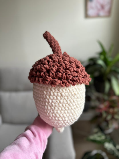 A crocheted acorn-shaped plush toy or decorative item held up by a person's hand. The top half is a dusty rose-brown textured crochet with a short stem, and the bottom half is cream/off-white.