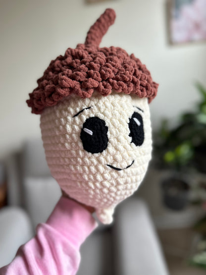 A crocheted acorn character plush toy held up by a person's hand. The acorn has a cream-colored face and body, large black embroidered eyes, a small stitched smile, and a textured brown acorn cap with a stem.