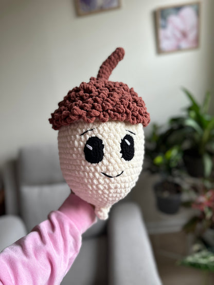 A handmade crochet acorn character toy with a smiling face, held up by a person wearing a pink sleeve in a living room setting.