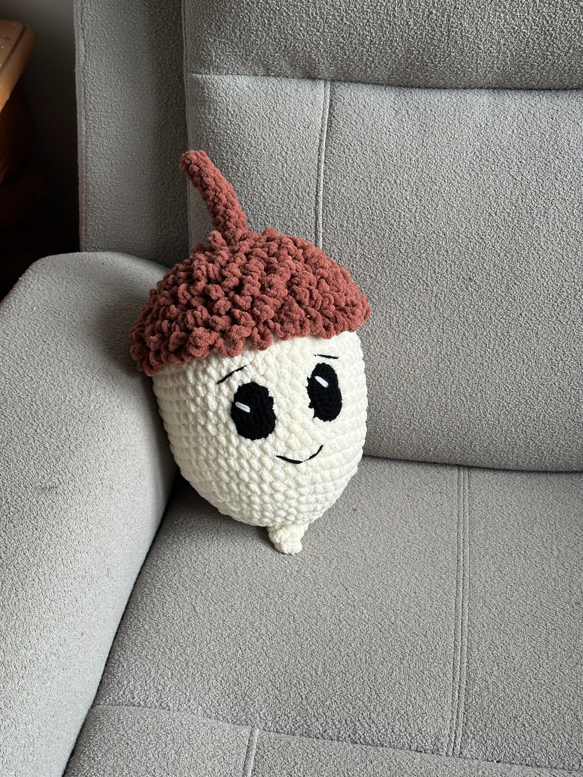 A crocheted acorn plush toy with a brown cap and stem, white body, and a smiling face, resting on a gray fabric armchair.