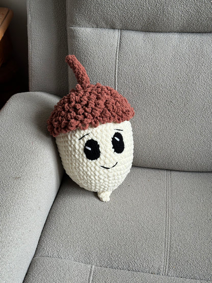A crocheted acorn plush toy with a brown cap and stem, white body, and a smiling face, resting on a gray fabric armchair.