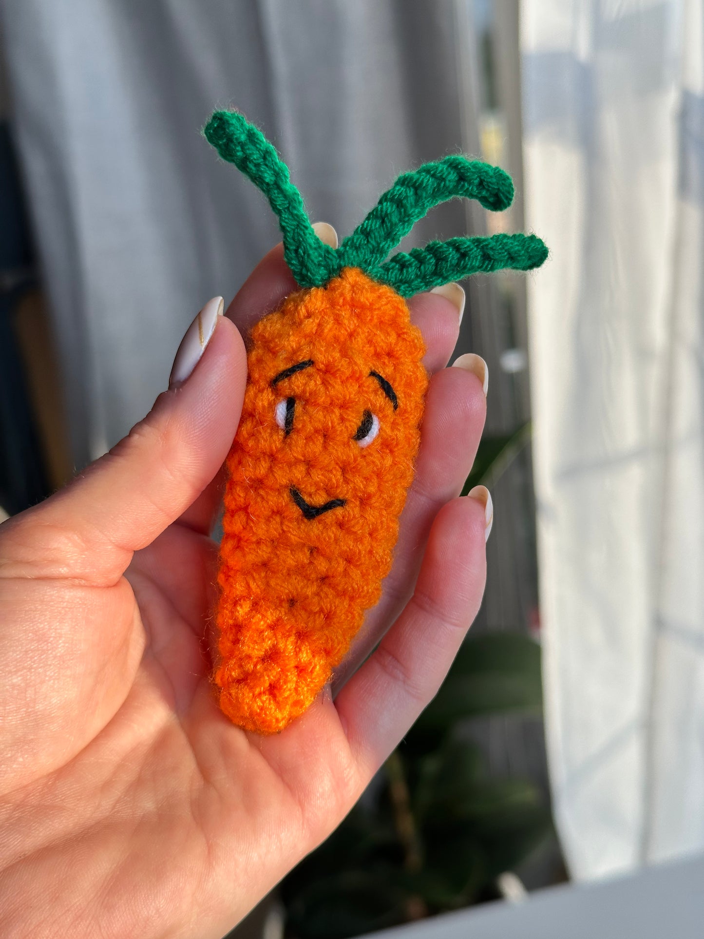 Close-up of crocheted carrot with embroidered face, held in hand near a bright window. Crochet Pattern Anti-stress carrot Clicker