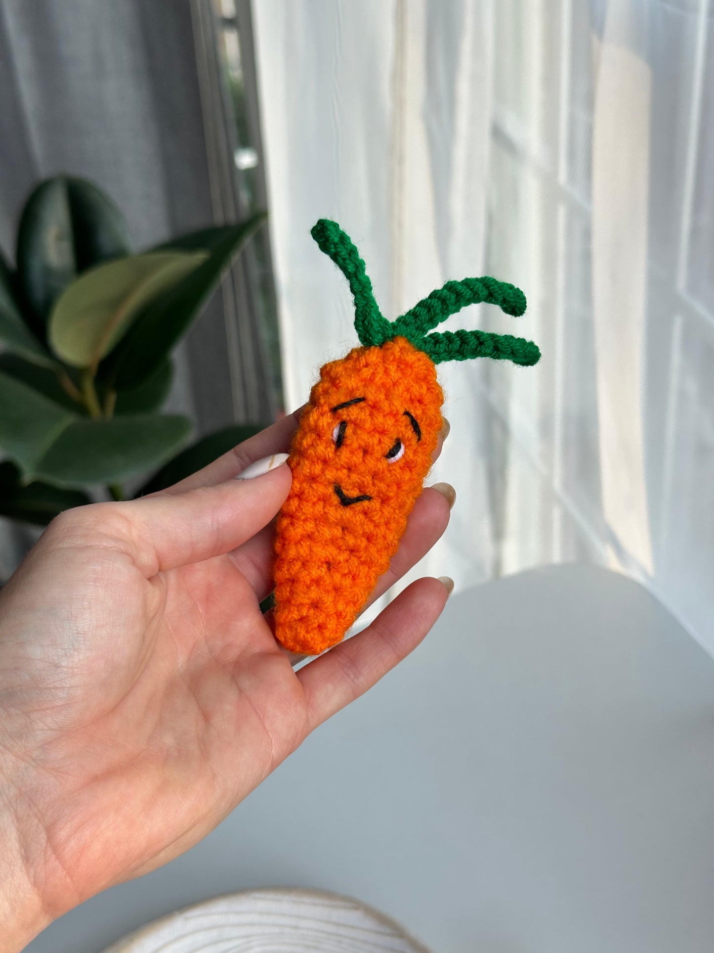 Crocheted carrot acrylic clicker anti-stress toy held in hand, positioned near white curtains with indoor plant in background.