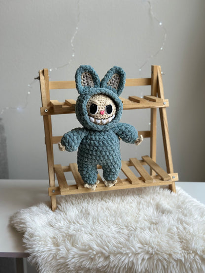 Crocheted Labubu made from light blue plushie yarn sitting on mini bench with cozy background. Crochet pattern