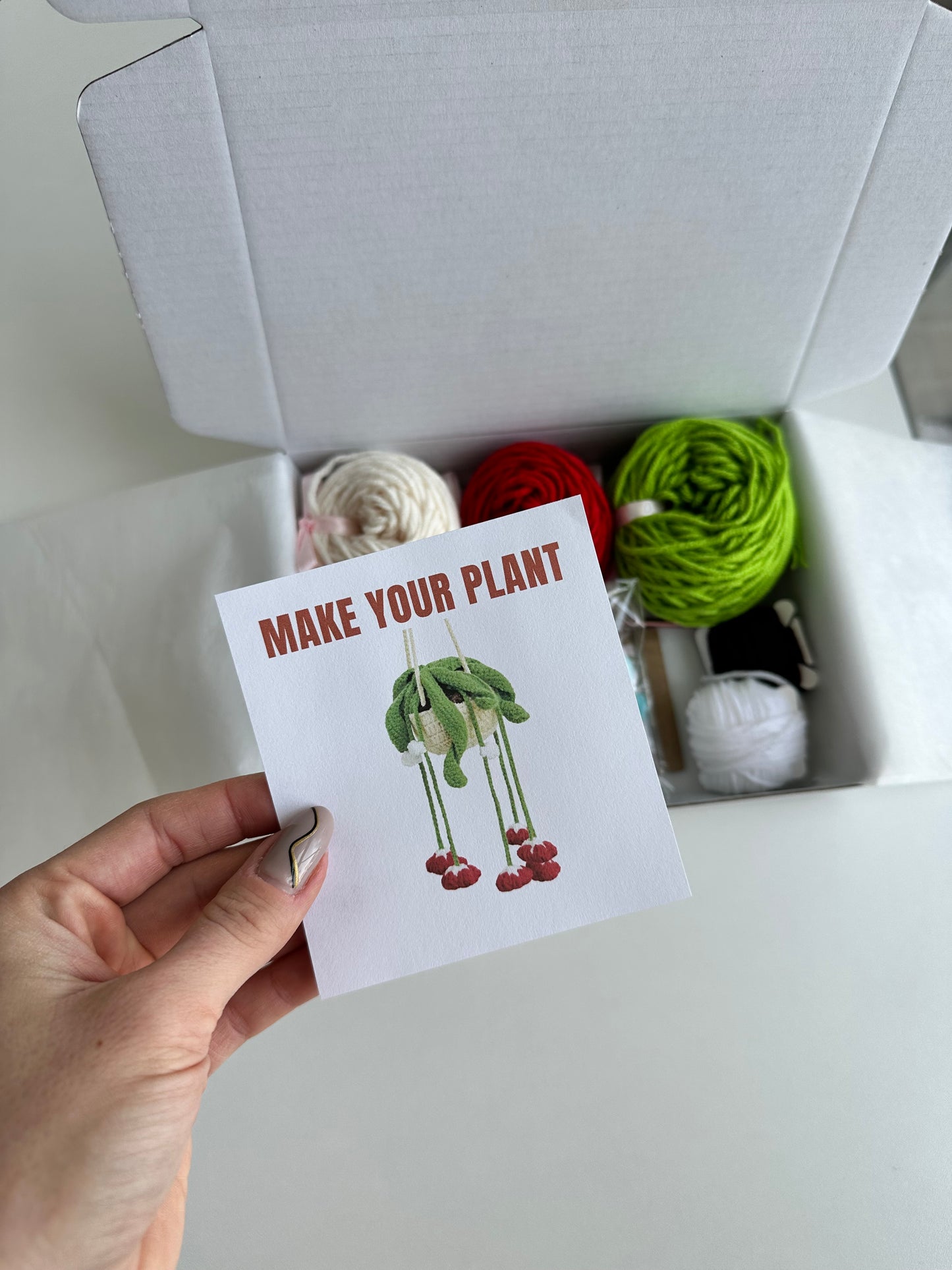 Open box held in hand, showing full set of materials from DIY Crochet Kit Red Bells Plant with branded card.