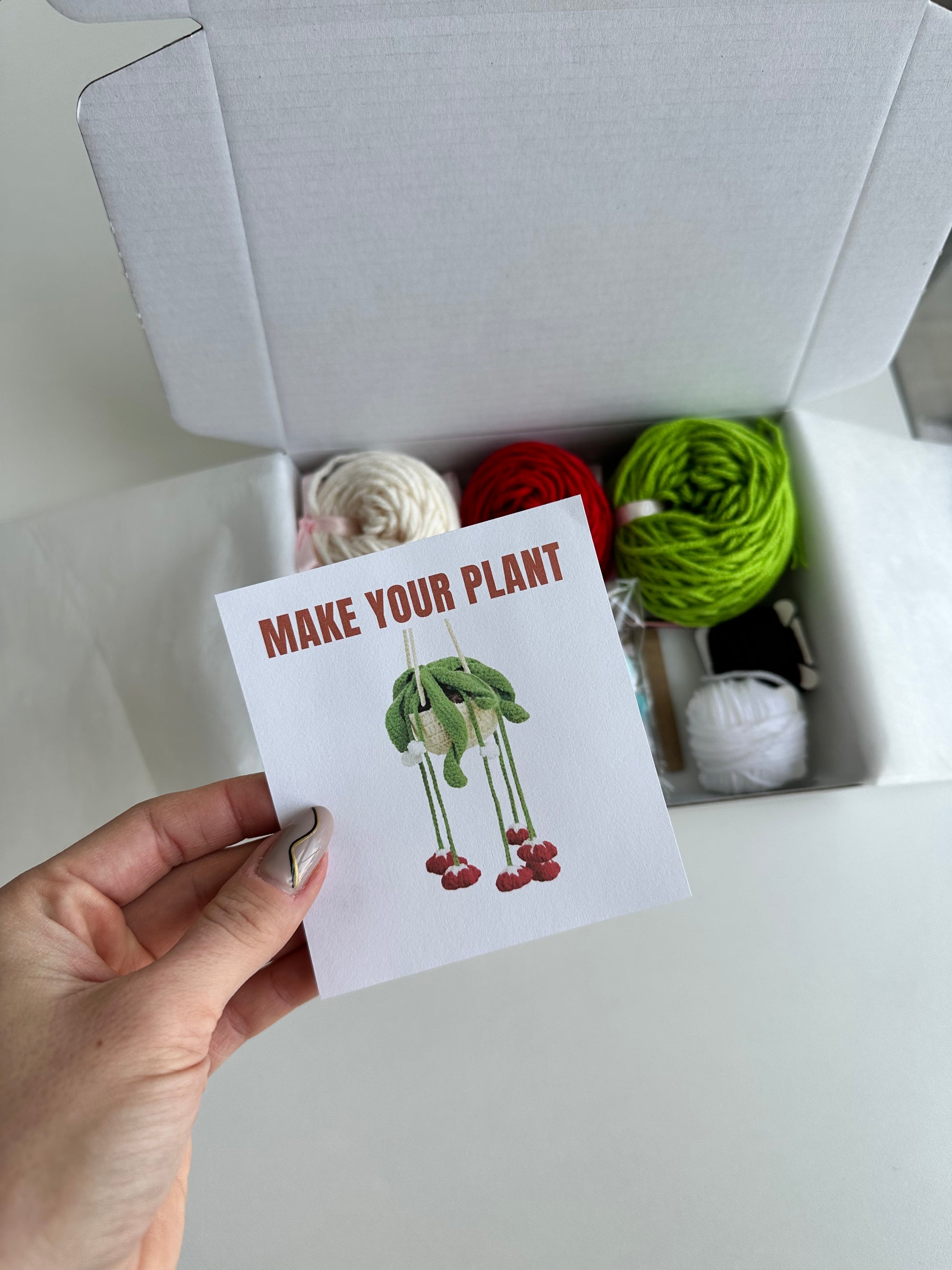 Open box held in hand, showing full set of materials from DIY Crochet Kit Red Bells Plant with branded card.