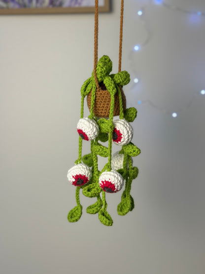 Completed spooky crochet plant with red and white eyeballs and green vines hanging against plain indoor wall. Crochet kit for Halloween