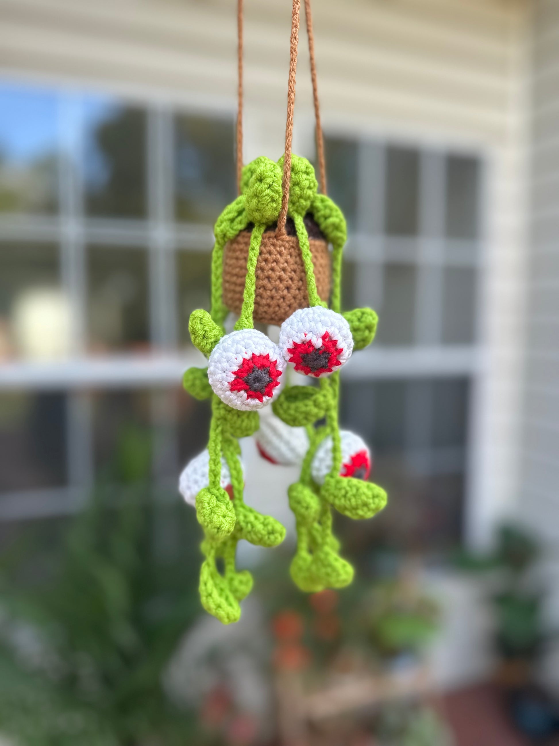 Crocheted decorative item with green leaves and red flowers hanging against a blurred outdoor background. Halloween Crochet Hanging Eyes pattern