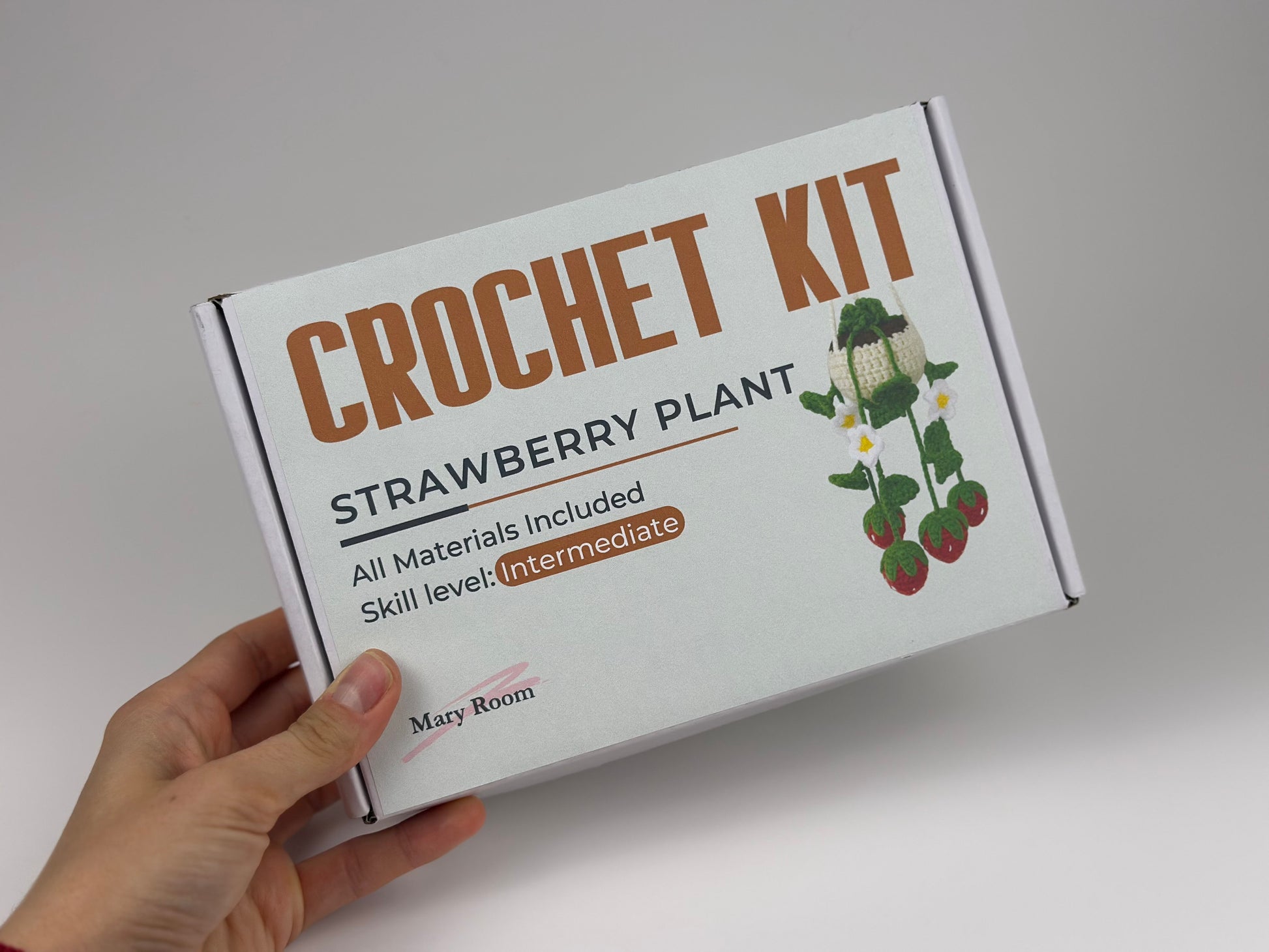 Crochet kit for strawberry plant held by a hand on a light gray background