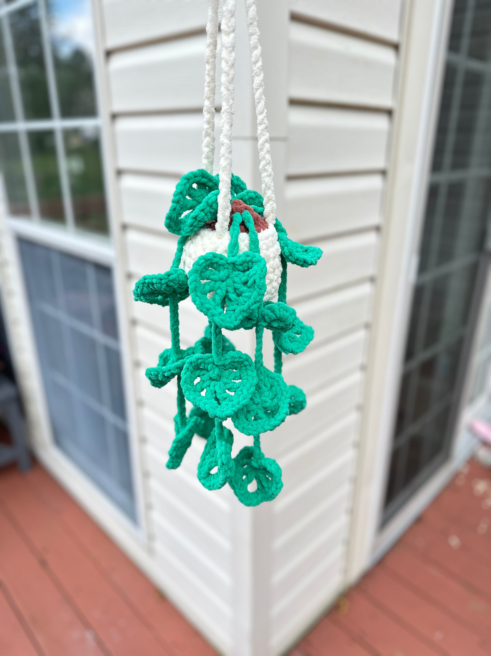Green crocheted Monstera plant decoration hanging from a white string against a window background.
