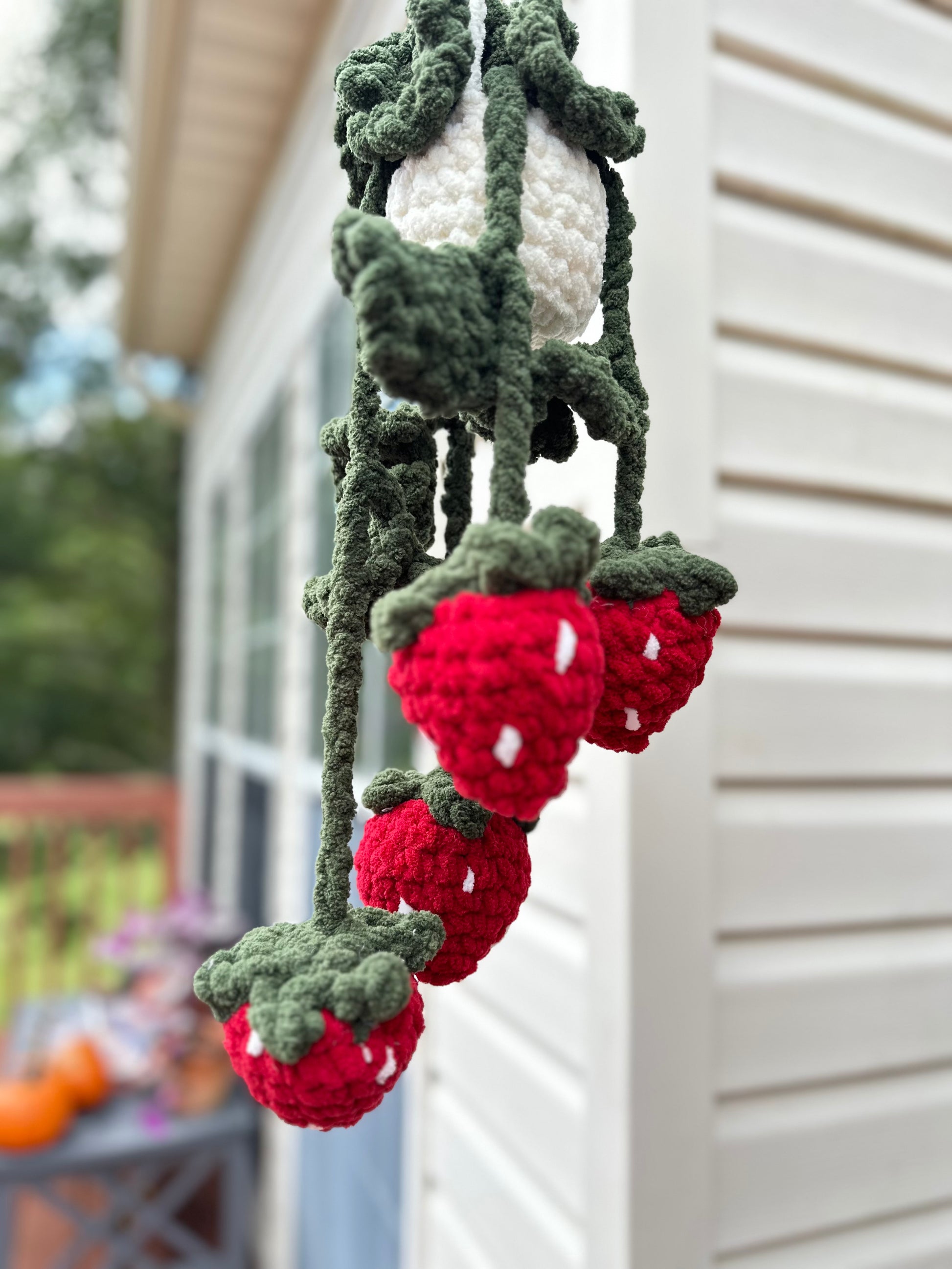 Crocheted strawberry plant with red strawberries and green leaves hanging against a house exterior.
