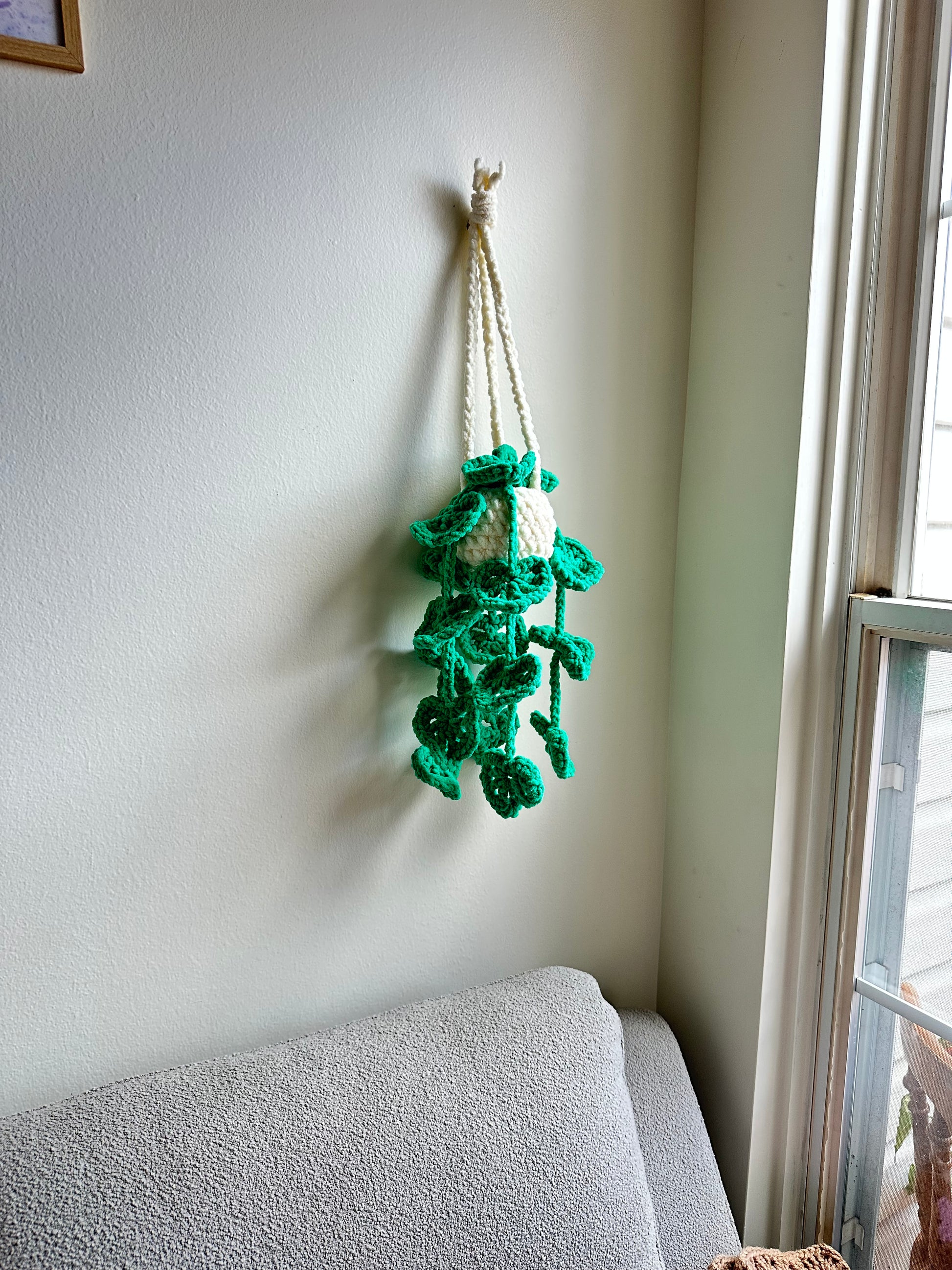 Green crocheted string of hearts plant decoration on a light-colored wall next to a window.