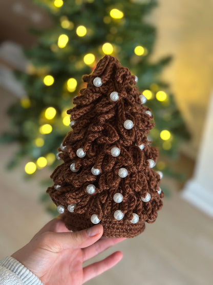 Hand holding a brown crocheted Christmas tree ornament with white beads against a blurred festive background.