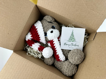 Handmade crochet Christmas teddy bear with red and white hat and scarf sitting in a gift box with Merry Christmas card