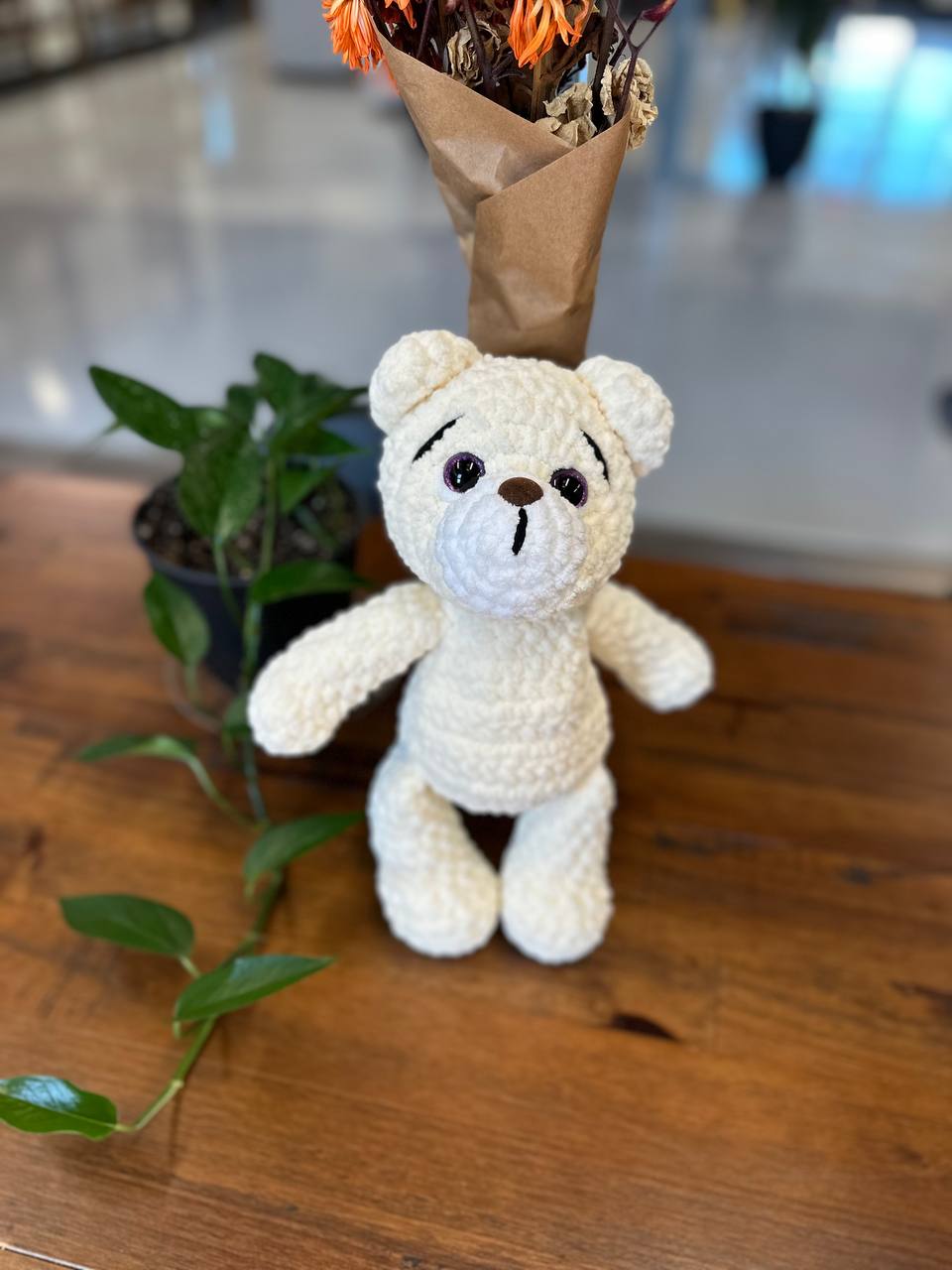 Cream crochet teddy bear displayed near a plant in cozy home decor style.