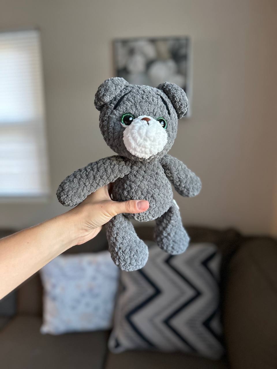 Grey crochet teddy bear shown on a couch in cozy home setting.