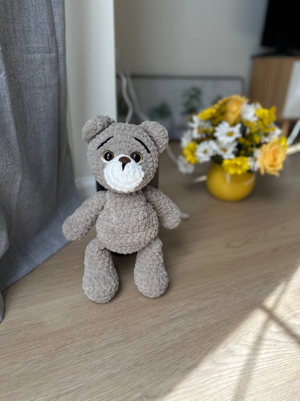 Grey plush teddy bear sitting near a window with yellow flowers in the background.