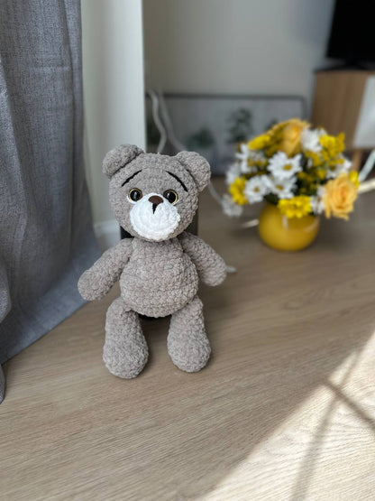 Grey plush teddy bear sitting near a window with yellow flowers in the background.