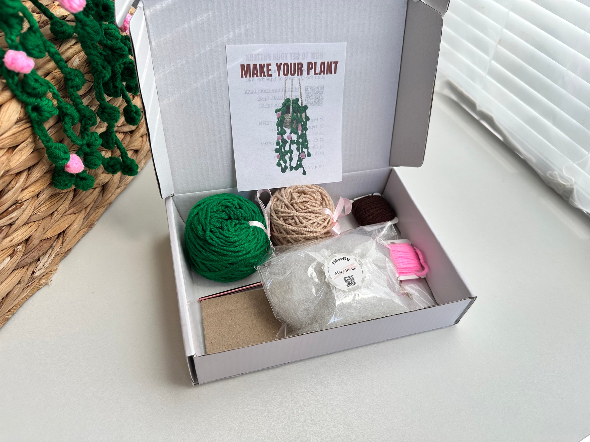 Box containing materials for making a hanging plant with a hook, needle, markers and yarn on a light surface.