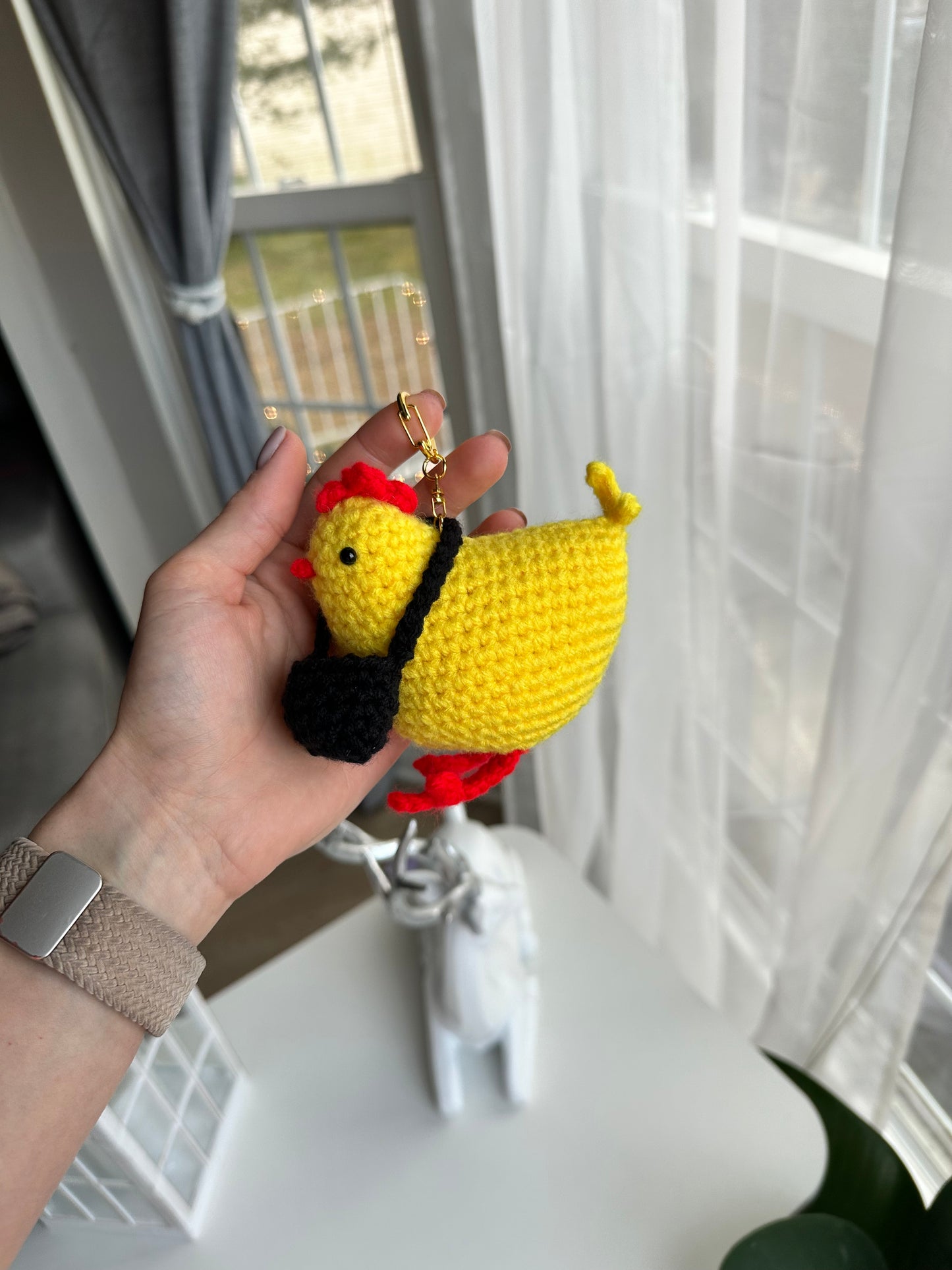 Hand holding a small yellow chicken keychain with a red bow in front of a window.