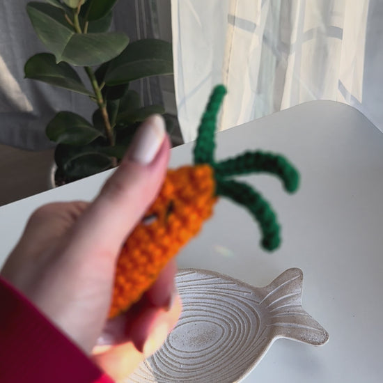 Hand holding crocheted carrot acrylic clicker anti-stress near a window with play button icon for crochet tutorial video.